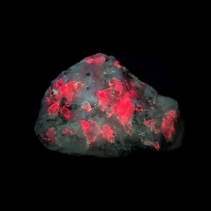 May include: A close-up of a mineral specimen with a dark background. The rock has a gray and white base color with bright red, irregular patches throughout. The red areas appear to glow, creating a striking contrast against the darker stone.