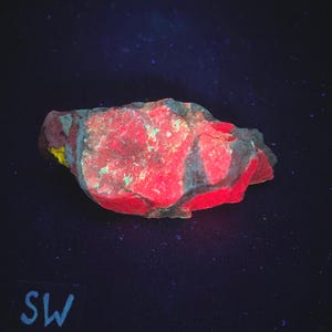 May include: A vibrant, irregularly shaped rock with a striking red hue, accented by patches of teal and yellow. The rock's surface has a rough texture, and the overall effect is enhanced by the dark background. The letters "SW" are visible in the lower left corner.