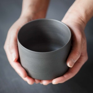 May include: A dark grey ceramic cup held in two hands.