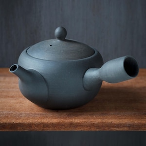 Może przedstawiać: Ciemnoszary ceramiczny czajnik z bocznym uchwytem i pokrywką z małym pokrętłem. Czajnik ma zaokrąglony korpus i dzióbek. Czajnik stoi na drewnianej powierzchni.