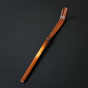 May include: A long, thin, wooden tea whisk with a curved end. The whisk is brown and has a natural wood finish.