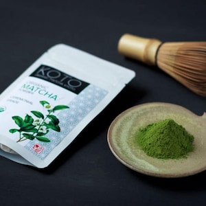 May include: A white pouch with the text "KOTO ORGANIC MATCHA CEREMONIAL GRADE" on it, a small bowl with green matcha powder, and a bamboo whisk.