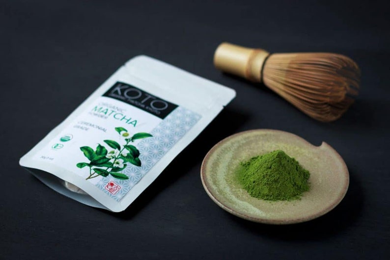 May include: A white pouch with the text "KOTO ORGANIC MATCHA CEREMONIAL GRADE" on it, a small bowl with green matcha powder, and a bamboo whisk.
