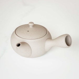 May include: A beige ceramic teapot with a round body, a lid with a small knob, and a side handle. The teapot has a spout and a speckled texture. The design is minimalist and suitable for brewing tea.