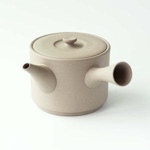 Op de afbeelding: Een beige keramische theepot met een rond lichaam en een deksel. De theepot heeft een tuit en een handvat dat aan de zijkant uitsteekt.