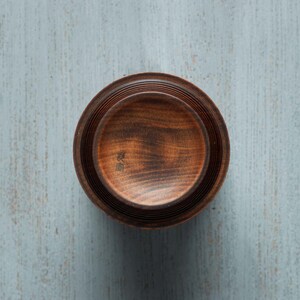 May include: A dark brown wooden bowl with a slightly raised rim and a smooth, polished finish. The bowl is made of multiple layers of wood, creating a unique and intricate design. The bowl has a small inscription in Japanese characters on the bottom.