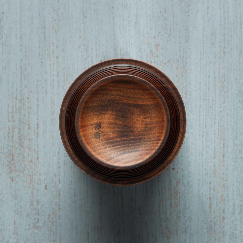 May include: A dark brown wooden bowl with a slightly raised rim and a smooth, polished finish. The bowl is made of multiple layers of wood, creating a unique and intricate design. The bowl has a small inscription in Japanese characters on the bottom.