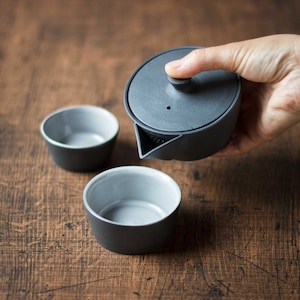 May include: A dark gray ceramic tea set with two small cups and a teapot with a built-in strainer. The teapot is being held by a hand. The cups and teapot have a matte finish and a light gray interior. The set is on a wooden surface.