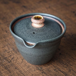 以下が含まれることがあります： 蓋付きの濃い灰色のセラミックティーポット。 蓋には、茶色と金の模様の小さな丸いハンドルが付いています。 ポットには注ぎ口があり、斑点のあるテクスチャです。 ポットの縁は赤茶色です。