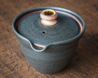 May include: A dark gray ceramic tea pot with a lid. The lid has a small, round handle with a brown and gold design. The pot has a spout and a speckled texture. The rim of the pot is a reddish-brown color.