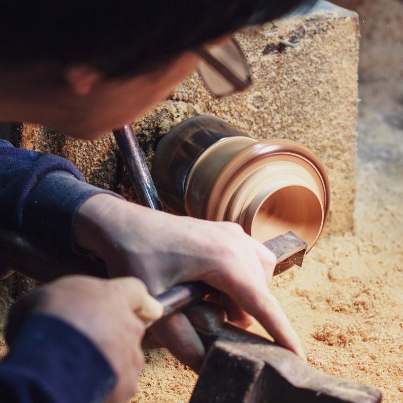 May include: A person using a woodturning lathe to shape a wooden bowl. The bowl is being turned on the lathe and the person is using a tool to shape the wood. The bowl is made of light brown wood and has a smooth, rounded surface.
