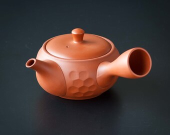 Może przedstawiać: Ceglastobrązowy ceramiczny czajnik z pokrywką i bocznym uchwytem. Czajnik ma okrągły korpus z wzorem plastra miodu z boku. Dzióbek jest mały i okrągły. Uchwyt to duża, otwarta rura.