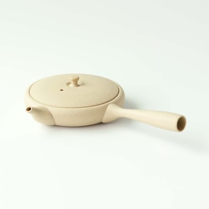 May include: A beige ceramic teapot with a round body and a spout. The teapot has a lid with a small knob on top. The teapot has a long handle.