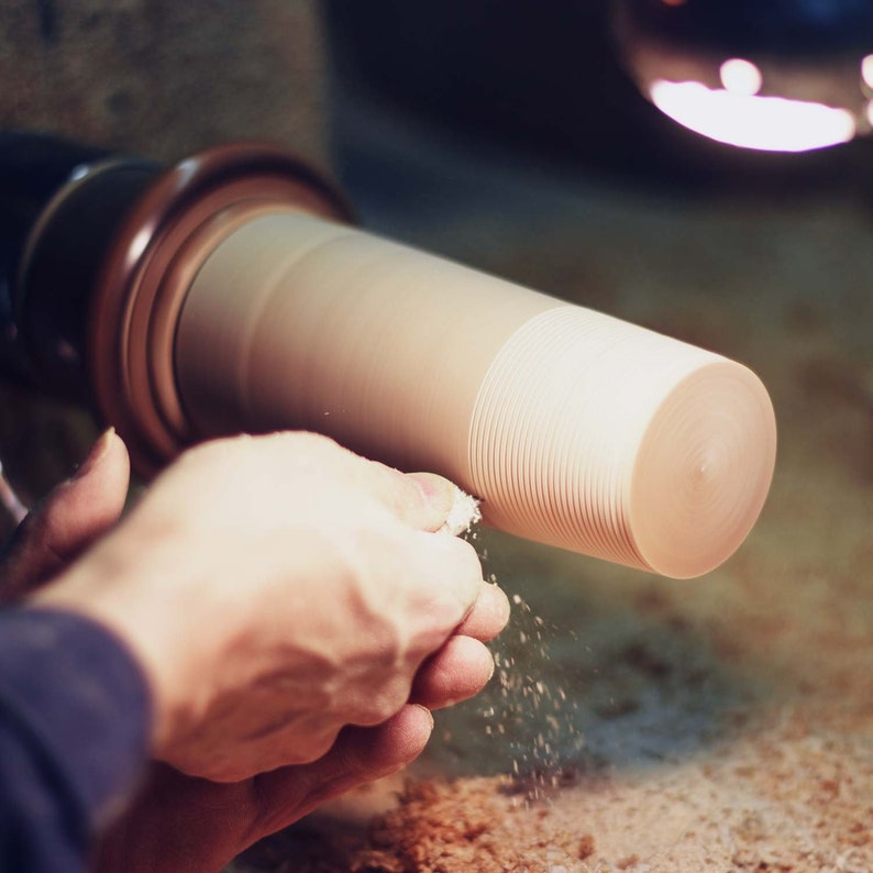May include: A close-up of a wooden object being turned on a lathe. The wood is light brown and has a smooth, even surface. Wood shavings are flying off the object as it spins.