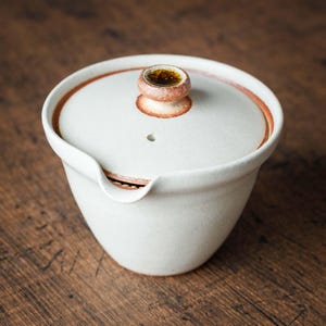 Pode incluir: Bule de chá de cerâmica branca com tampa marrom e uma pequena pega marrom no topo. A chaleira tem um pequeno bico lateral.