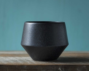 Puede incluir: Una taza de cerámica negra con forma geométrica. La taza está sobre una superficie de madera.