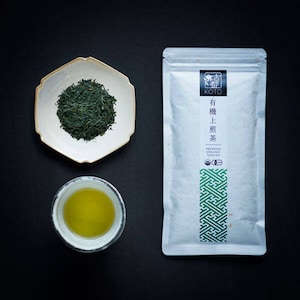 Könnte beinhalten: Eine weiße Packung Bio-Grüntee der Marke Koto mit japanischem Text, der "Koto, Bio-Sencha, Premium" lautet, und einem grünen geometrischen Muster. Außerdem sind eine kleine weiße Untertasse mit grünen Teeblättern und eine weiße Tasse mit grünem Tee zu sehen.