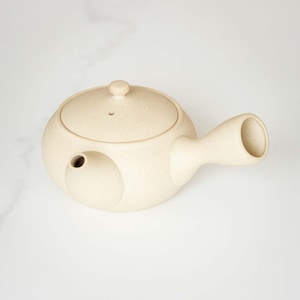 Può includere: Una teiera in ceramica beige con un corpo arrotondato e un beccuccio laterale. La teiera ha un coperchio con un piccolo pomo in cima e un lungo manico curvo.