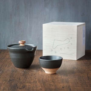 Puede incluir: Un juego de tetera y taza de té de cerámica negra con una caja de madera. La caja tiene el texto "A Piece" impreso en ella.