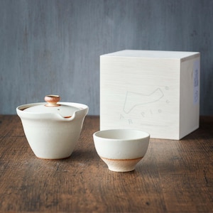 Puede incluir: Un juego de té de cerámica blanca, que incluye una tetera con tapa y una taza pequeña. También hay una caja de madera con la palabra "APPLE" y un diseño gráfico. El juego está colocado sobre una superficie de madera.