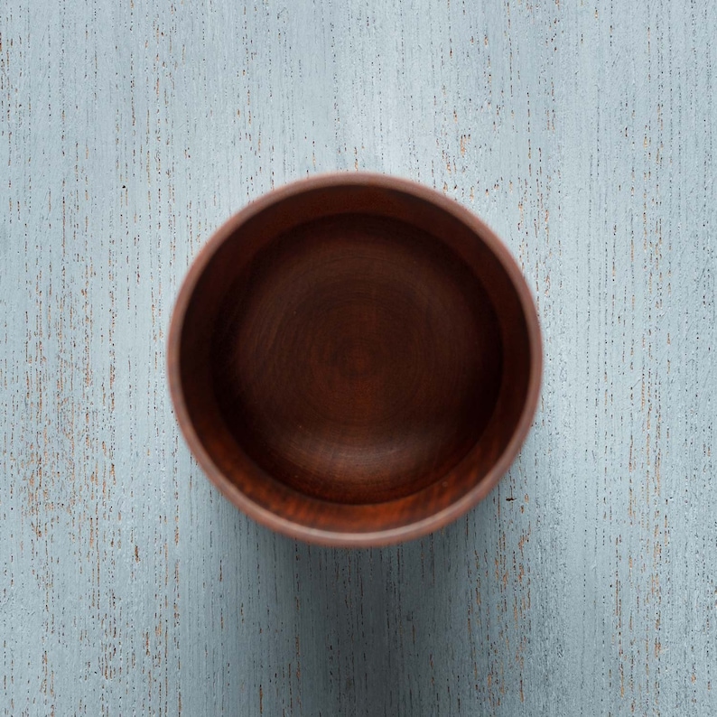 May include: A dark brown wooden bowl with a smooth, rounded interior. The bowl is empty and sits on a light blue wooden surface.