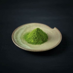 May include: A small pile of bright green matcha powder sits in a shallow, light brown ceramic dish. The dish has a textured surface and a slightly raised rim.