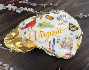 May include: A decorative oyster shell with a gold rim, featuring a colourful Virginia-themed design. The shell includes a red cardinal, the word "Virginia", and illustrations of a lighthouse, ship, and other state symbols.