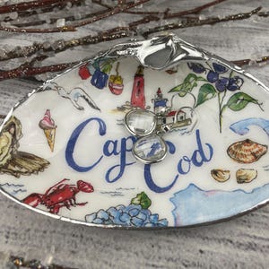 May include: A decorative seashell trinket dish with a hand-painted coastal design. The dish features a lighthouse, lobster, ice cream cone, and the words "Cap Cod" in blue script. A pair of clear earrings rests inside.