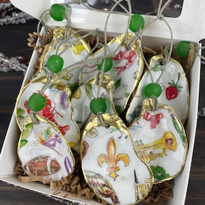 May include: A set of hand-painted oyster shell ornaments in a white box. Each shell features unique designs with gold edging, including a football, fleur-de-lis, and a trumpet. Green beads and twine are attached for hanging.