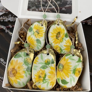 May include: A set of six decorative ornaments in a white box. Each ornament is shell-shaped with a gold-painted edge and a sunflower design. They have a string for hanging and a small green bead.