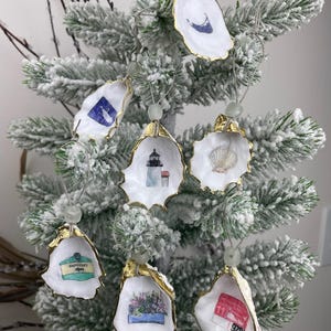 May include: A collection of seven oyster shell ornaments with gold-painted edges, each featuring unique illustrations including a lighthouse, a seashell, a floral arrangement, and a Nantucket sign, suspended by strings with small light green beads.