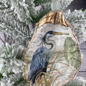 May include: An oyster shell Christmas ornament featuring a blue heron illustration. The shell has a gold-painted edge and is decorated with a map design. The ornament hangs from a string with a gold bead and a light blue glass bead.