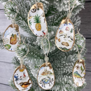 May include: Hand-painted oyster shell ornaments with gold edges, each featuring a unique coastal design. The ornaments depict images of a pineapple, crab, sailboat, and other beach-themed illustrations. They are strung with twine and a small bead for hanging on a Christmas tree.