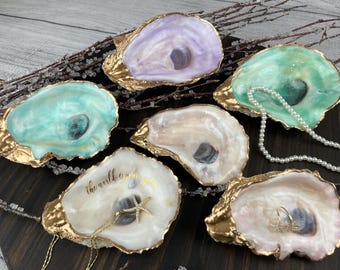 May include: Six decorative oyster shell dishes with gold-painted edges. The shells are painted in various colours, including light blue, green, and lavender. One shell has the text "the world is your oyster" in gold script. Some shells contain jewellery.