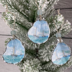 Könnte beinhalten: Drei handgefertigte Austernschalen-Ornamente mit nautischem Thema. Jede Schale zeigt ein Aquarellgemälde von Segelbooten auf blauem Meer unter blauem Himmel. Die Schalen sind silberfarben eingefasst und mit Schnur und einer blauen Perle aufgehängt.