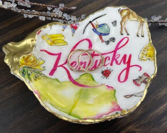 May include: A hand-painted oyster shell ring dish with gold trim. The dish features the word "Kentucky" in pink script, along with watercolor illustrations of state symbols and two gold rings. The background is white.