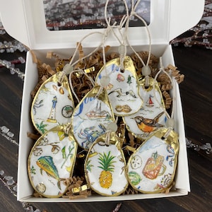 May include: A set of hand-painted oyster shell ornaments with gold trim, each featuring a unique coastal design. The designs include a lighthouse, sailboat, crab, pineapple, and a bird. The ornaments are displayed in a white box with decorative filler.