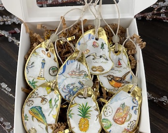 May include: A set of hand-painted oyster shell ornaments with gold trim, each featuring a unique coastal design. The designs include a lighthouse, sailboat, crab, pineapple, and a bird. The ornaments are displayed in a white box with decorative filler.