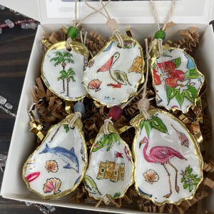 May include: A set of six hand-painted oyster shell ornaments. Each shell features a unique design, including a palm tree, bird, flowers, dolphin, castle, and flamingo. The shells have gold-painted edges and are strung with twine and beads.