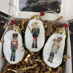 Puede incluir: Tres adornos navideños de concha de ostra con ribete dorado y colgadores de cuerda. Cada concha presenta una ilustración diferente de un personaje de Cascanueces. Los adornos se exhiben en una caja blanca con relleno dorado y marrón.