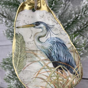 May include: An oyster shell ornament featuring a blue heron illustration. The shell has a gold-painted edge and a natural twine hanger. The heron is depicted in a marsh setting with the word "OCEAN" visible. The ornament is hanging on a snowy evergreen branch.