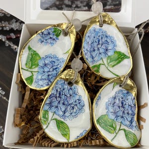 May include: Four oyster shell ornaments with gold trim and blue hydrangea flower designs. The ornaments are packaged in a white box with brown paper filler.