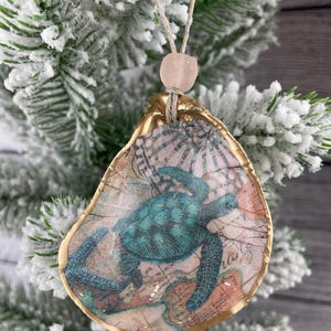 May include: A decorative oyster shell ornament featuring a decoupage print of a teal sea turtle over a vintage map background. The shell has a metallic gold painted edge and is suspended by a tan twine string with a single pale pink bead.