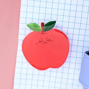 May include: A red apple sticker with a cute cartoon face. The apple has a green stem and leaves. The sticker is on a white gridded paper background.