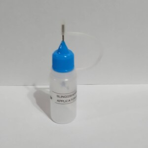 May include: A clear plastic applicator bottle with a blue cap and a thin metal applicator tip. The bottle has white text that reads "BLINGSHOP APPLICATOR". The bottle is filled with a white liquid.