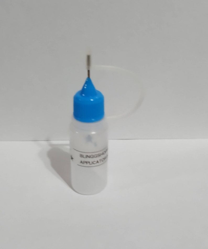 May include: A clear plastic applicator bottle with a blue cap and a thin metal applicator tip. The bottle has white text that reads "BLINGSHOP APPLICATOR". The bottle is filled with a white liquid.