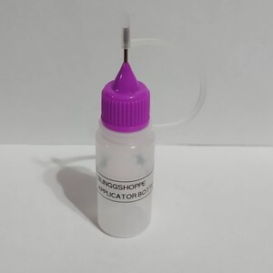 May include: A clear plastic applicator bottle with a purple cap and a thin metal needle. The bottle has a label that reads "BLINGGSHOPPE APPLICATOR BOTTLE". The cap is ribbed, and a clear plastic tube extends from the bottle.