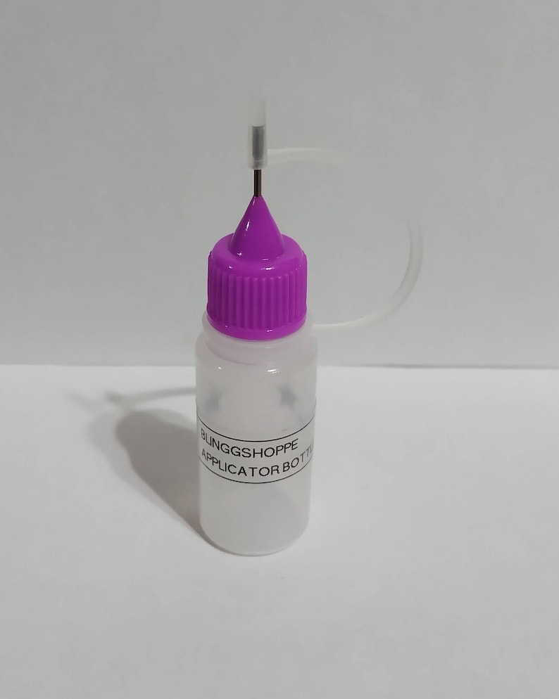 May include: A clear plastic applicator bottle with a purple cap and a thin metal needle. The bottle has a label that reads "BLINGGSHOPPE APPLICATOR BOTTLE". The cap is ribbed, and a clear plastic tube extends from the bottle.