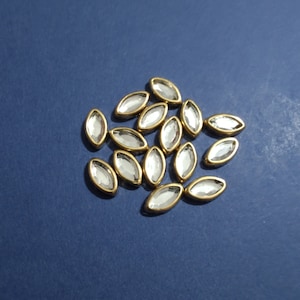 May include: A group of clear glass oval-shaped rhinestones with gold settings. The rhinestones are scattered on a blue background.