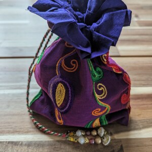 May include: A purple drawstring bag with colorful embroidery and a blue fabric top. The bag has a tassel with beads.